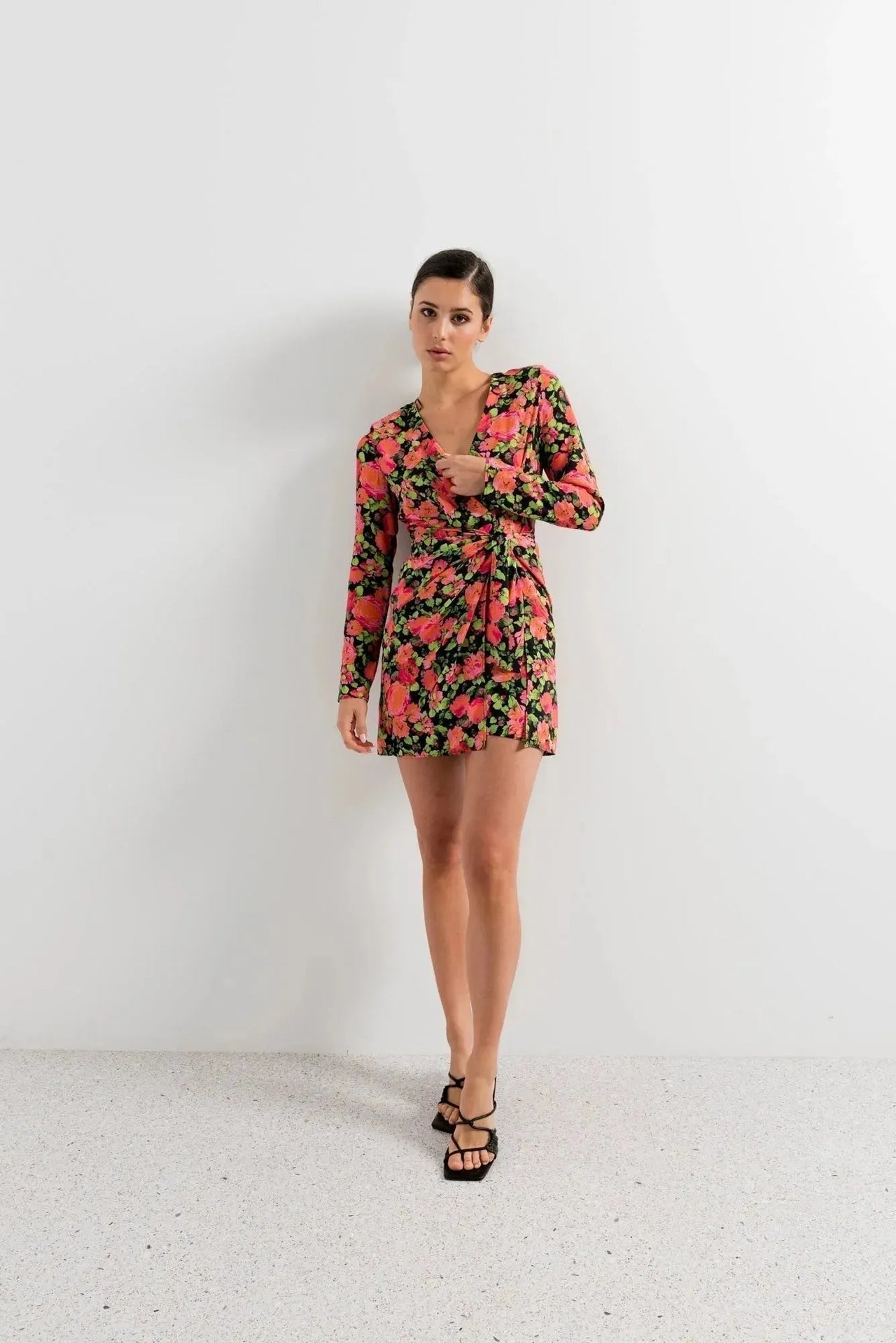 Femme en position posée montrant sa robe portefeuille femme fleuri noir et rose, courte à manches longues avec décolleté en V, parfaite pour diverses occasions.