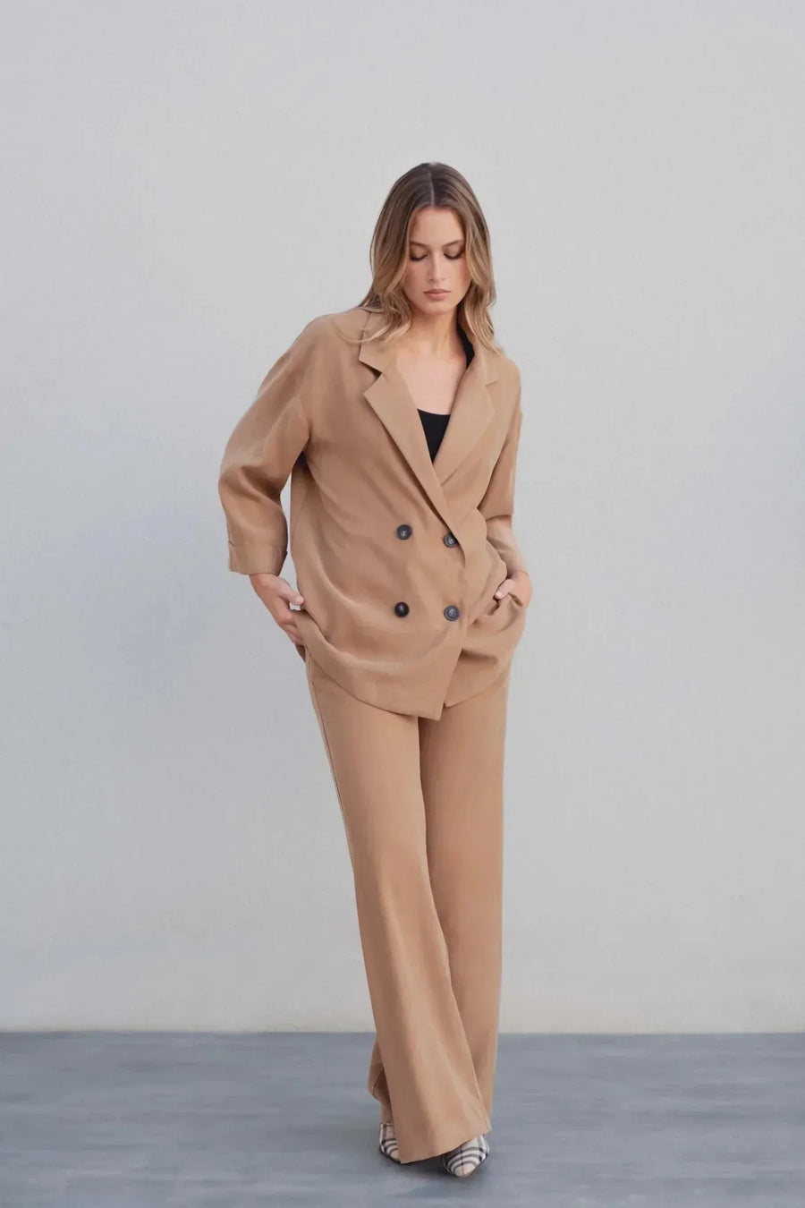 Femme debout en tailleur blazer fluide camel avec un pantalon assorti, posant avec élégance.