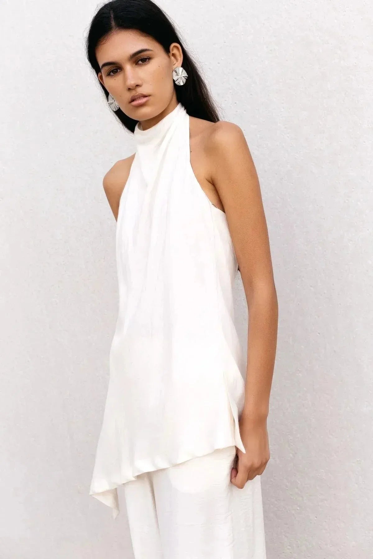 Haut dos nu femme chic Ivoire à col montant halter, coupe asymétrique élégante, porté par une mannequin avec de grands accessoires, sur fond neutre.