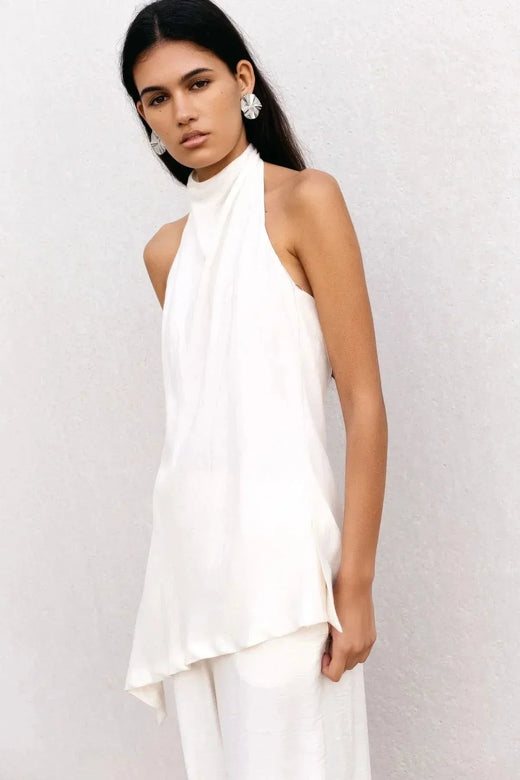 Haut dos nu femme chic Ivoire à col montant halter, coupe asymétrique élégante, porté par une mannequin avec de grands accessoires, sur fond neutre.