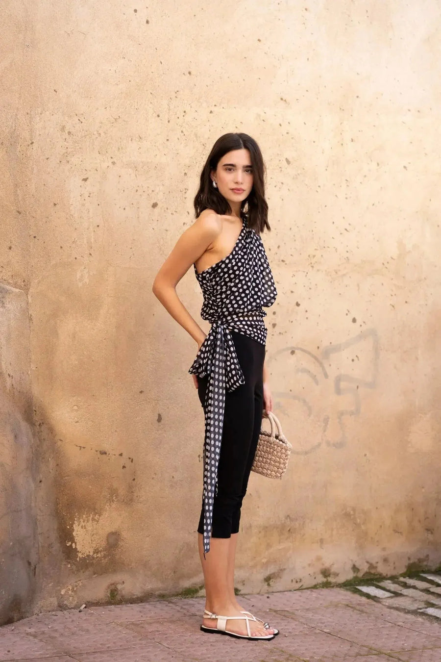 Femme portant un top asymétrique imprimé à pois noirs et blancs, pantalon noir et sandales blanches