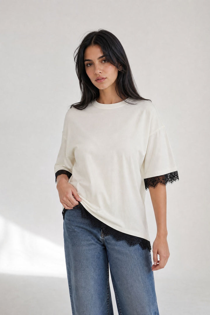 Top oversize crème à dentelle noire porté de trois quarts, finitions aux manches et à l’ourlet asymétrique.