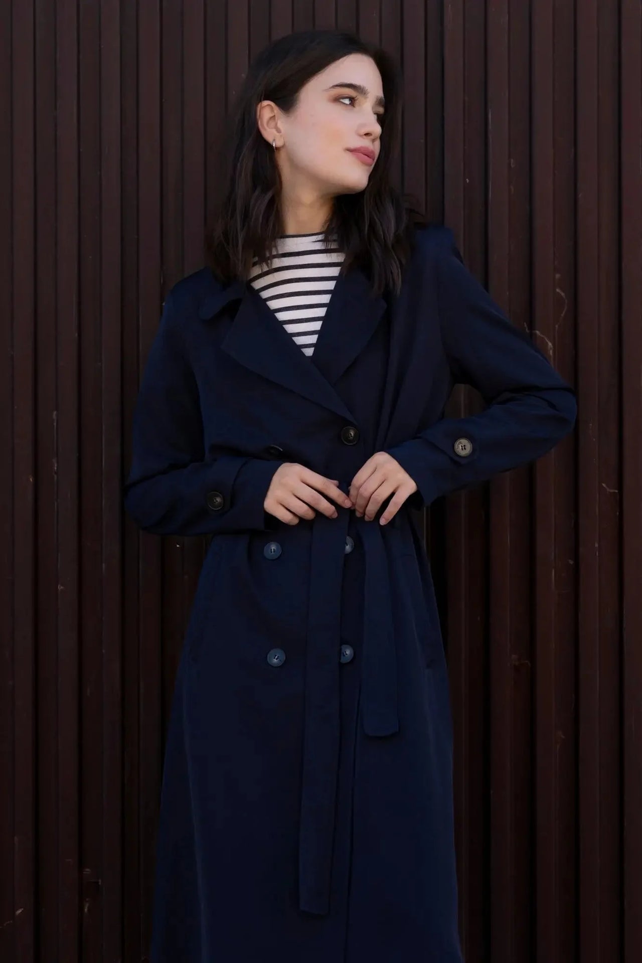 Femme dans un trench coat femme ample long ceinturé, avec une expression sereine, face à une mur en bois.