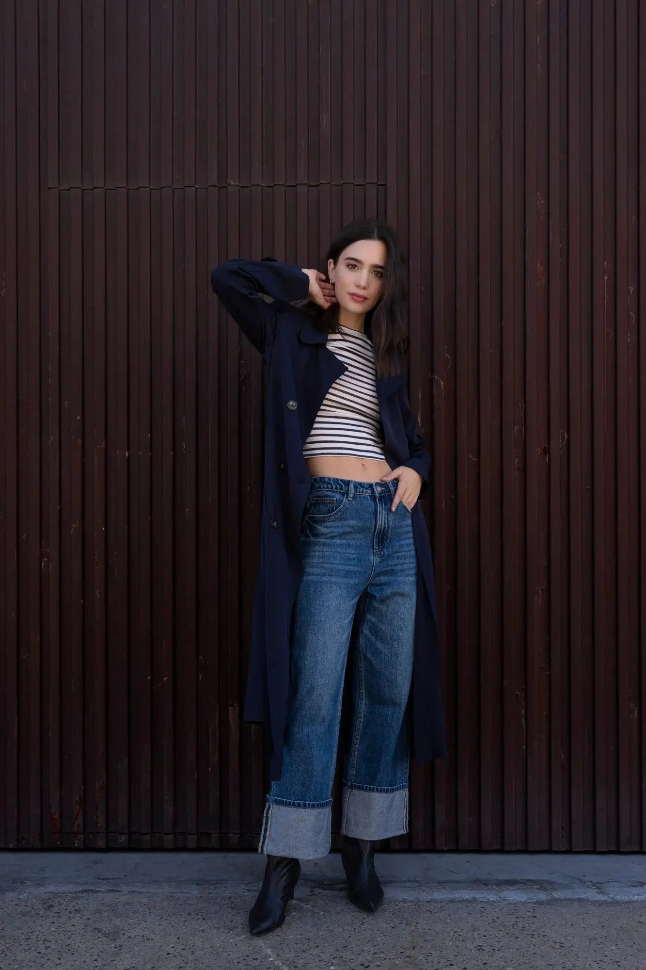 Femme portant un trench-coat ample avec ceinture, crop top rayé, jeans retroussé et bottes noires