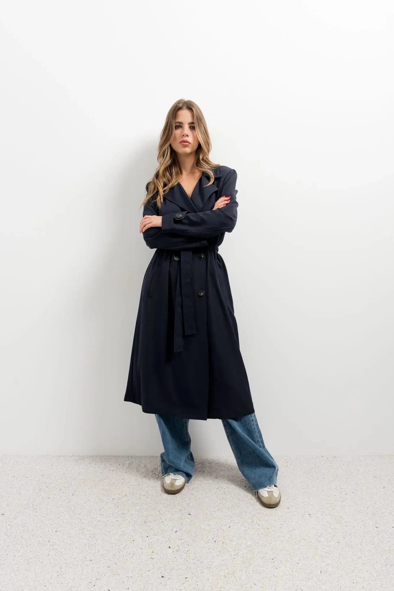 Femme portant un trench-coat ample avec ceinture, jeans bleus et sneakers clairs
