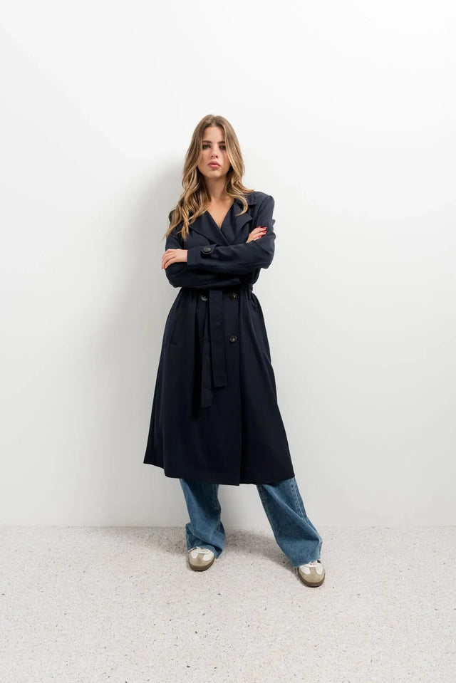 Femme stylée dans un trench coat femme ample long ceinturé, se tenant en pose décontractée avec un air confiant.